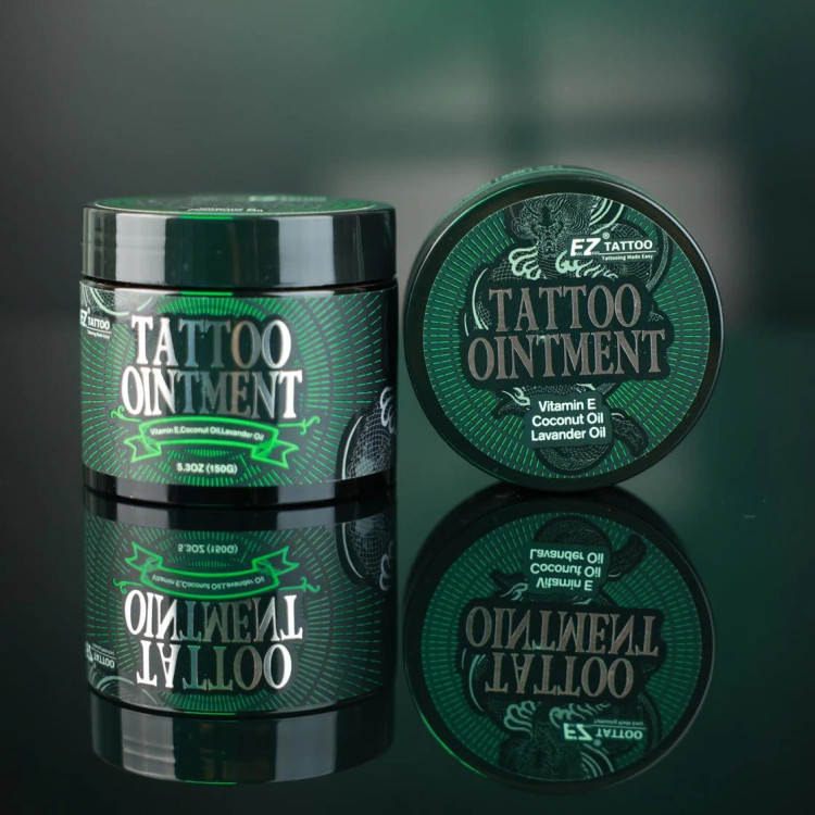 نمای تبلیغاتی کرم حین کار تتو EZ Tattoo وزن ۱۵۰ گرم، نمایش قوطی و درب محصول روی سطح براق با انعکاس تصویر، حاوی ویتامین E، روغن نارگیل و روغن اسطوخودوس