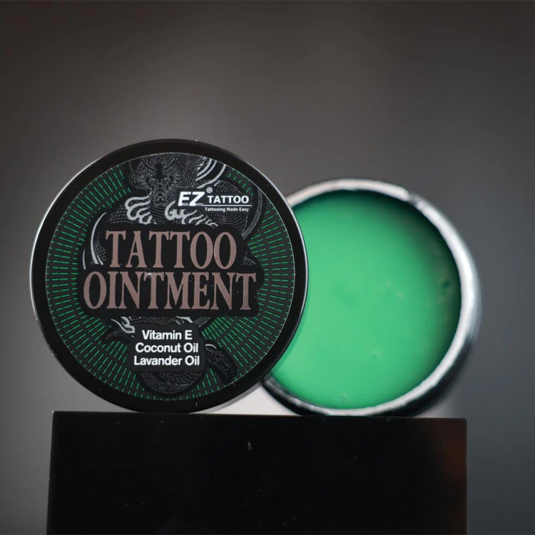 نمای باز کرم حین کار EZ Tattoo 150 گرم، نمایش بافت سبز و نرم محصول در کنار درب قوطی، حاوی ویتامین E، روغن نارگیل و روغن اسطوخودوس، مناسب مرطوبسازی پوست و استفاده حین انجام تتو